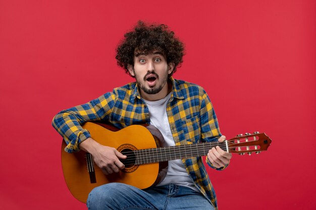 Vorderansicht junger Mann, der auf der roten Wand sitzt und Gitarre spielt, Live-Farbbandmusik spielt Applaus