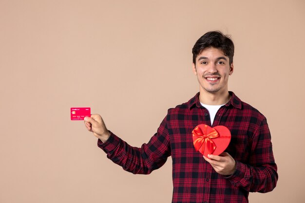 Vorderansicht junger Mann, der herzförmiges Geschenk und Bankkarte an brauner Wand hält