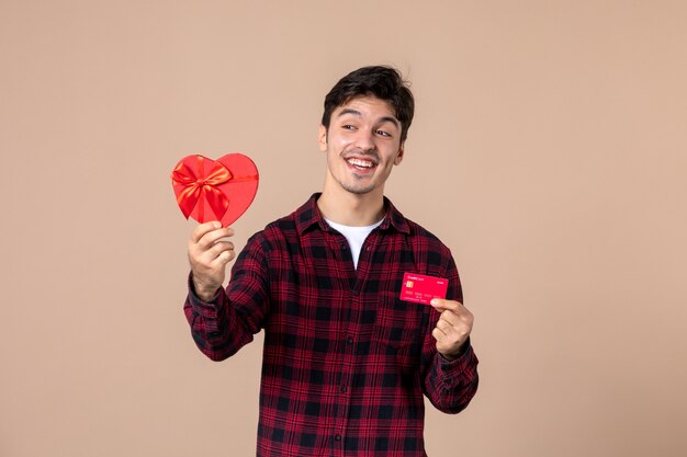 Vorderansicht junger Mann, der herzförmiges Geschenk und Bankkarte an brauner Wand hält