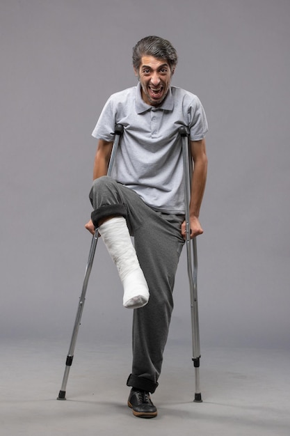 Vorderansicht junger Mann, der Krücken aufgrund eines gebrochenen Fußes an einer grauen Wand verwendet, deaktiviert den gebrochenen Unfallbeinfuß