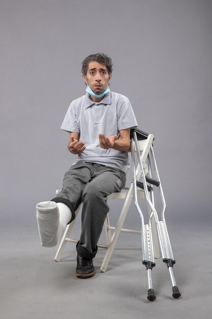Vorderansicht junger Mann, der mit bandagiertem gebrochenem Fuß auf dem grauen Wandschmerz sitzt