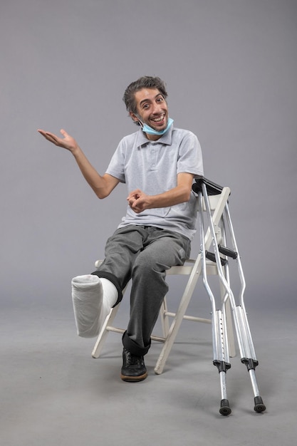 Vorderansicht junger Mann, der mit bandagiertem gebrochenem Fuß auf grauem Wandschmerz Beinunfall männlicher Fußdrehung sitzt
