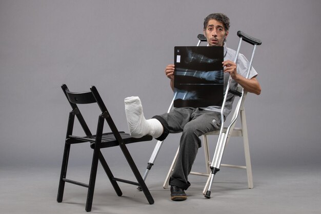Vorderansicht junger Mann, der mit gebrochenem Fuß mit Krücken sitzt und Röntgen auf der grauen Wand hält, Fußdrehung gebrochener Schmerzunfall