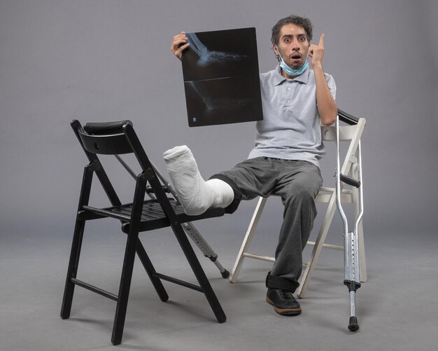 Vorderansicht junger Mann, der mit gebrochenem Fuß sitzt und ein Röntgenbild davon auf der grauen Wand hält
