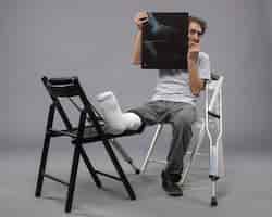 Kostenloses Foto vorderansicht junger mann, der mit gebrochenem fuß sitzt und ein röntgenbild davon auf der grauen wand hält