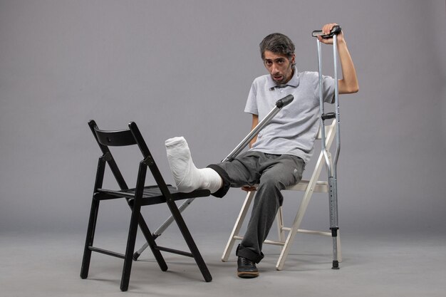 Vorderansicht junger Mann, der mit gebrochenem Fuß sitzt und Krücken auf grauem Boden verwendet