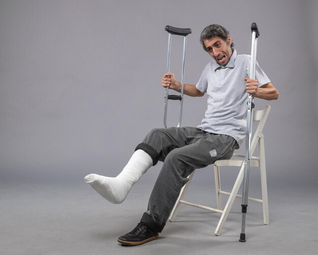 Vorderansicht junger Mann, der mit gebrochenem Fuß sitzt und Krücken auf grauem Wandfuß hält