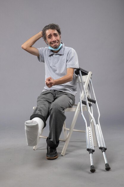 Vorderansicht junger Mann, der mit gebrochenem Fuß und gebundenem Verband auf der grauen Wand sitzt