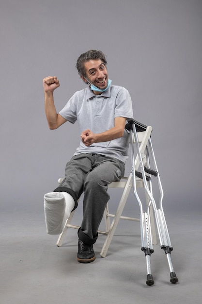 Vorderansicht junger Mann, der mit gebrochenem Fuß und gebundenem Verband auf grauem Wandschmerz-Unfallfußdrehbeinmann sitzt