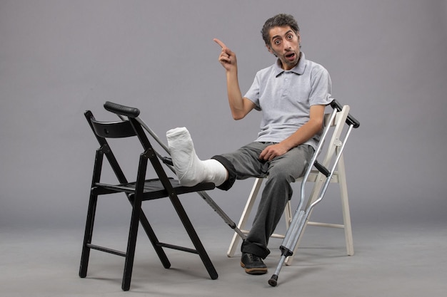 Vorderansicht junger Mann, der mit gebrochenem Fuß und Krücken auf grauen Wandunfallbeinen sitzt