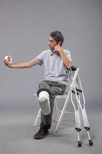 Vorderansicht junger Mann, der mit gebrochenem Fußverband sitzt und Uhr an der grauen Wand hält