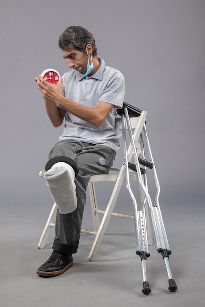 Vorderansicht junger Mann, der mit gebundenem Verband aufgrund eines gebrochenen Fußes sitzt und die Uhr an der grauen Wand hält, verdrehen Schmerzen, Unfallbein, männlicher Fuß