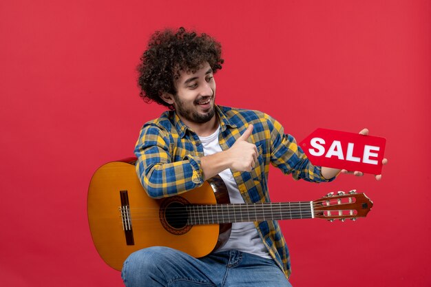 Vorderansicht junger Mann, der mit Gitarre auf roter Wand sitzt Live-Musik-Farbapplaus-Musiker-Verkaufsspiel