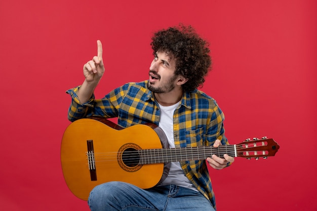 Vorderansicht junger Mann, der mit Gitarre auf roter Wand sitzt und Musik-Performance-Musiker-Farb-Live-Konzert spielt