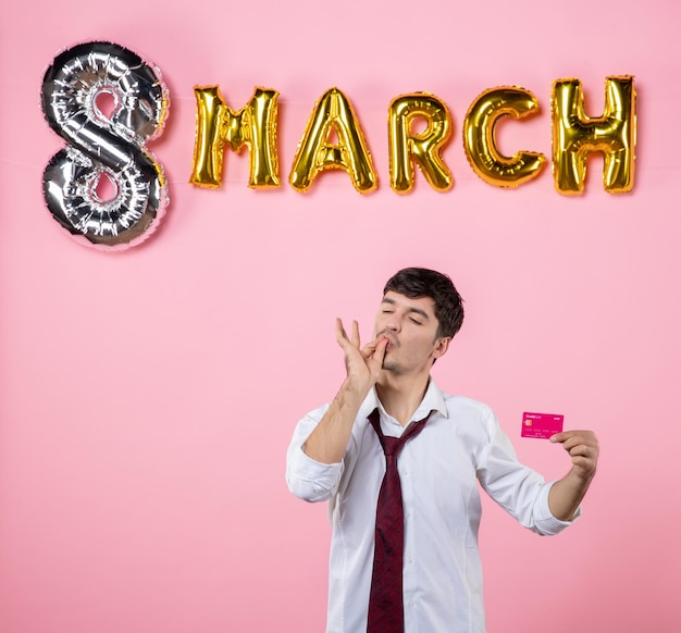 Kostenloses Foto vorderansicht junger mann, der rosa bankkarte mit marschdekoration auf rosa hintergrund hält partygeschenk urlaub mann geld gleichheit farbe einkaufen