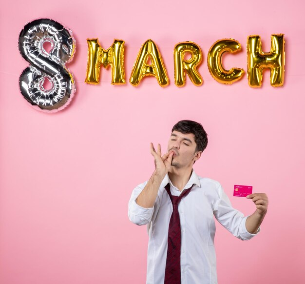 Vorderansicht junger Mann, der rosa Bankkarte mit Marschdekoration auf rosa Hintergrund hält Partygeschenk Urlaub Mann Geld Gleichheit Farbe einkaufen