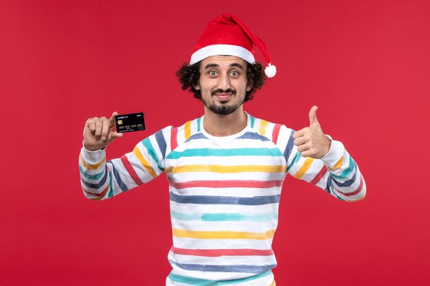 Vorderansicht junger Mann, der schwarze Bankkarte auf roten Neujahrsgeldferien der roten Wand hält
