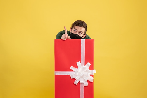 Vorderansicht junger Mann, der sich hinter großer Geschenkbox auf Gelb versteckt