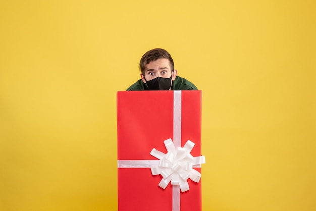 Vorderansicht junger Mann, der sich hinter großer Geschenkbox auf Gelb versteckt