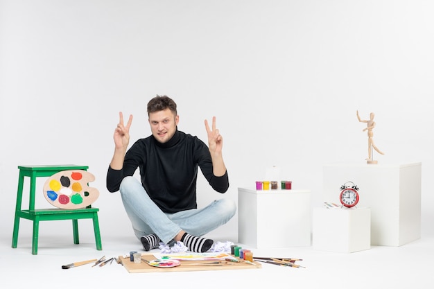 Vorderansicht junger Mann, der um Farben und Quasten zum Zeichnen auf weißer Wandkunst sitzt, zeichnet Farbbildkünstler malt Malerei