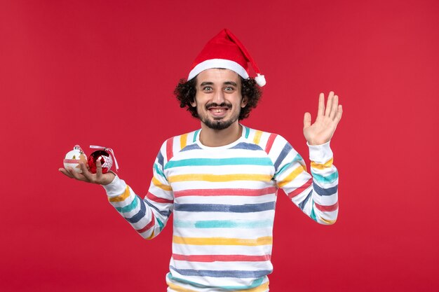 Vorderansicht junger Mann, der Weihnachtsbaumspielzeug auf menschlichem Feiertagsrot der roten Wand des neuen Jahres hält