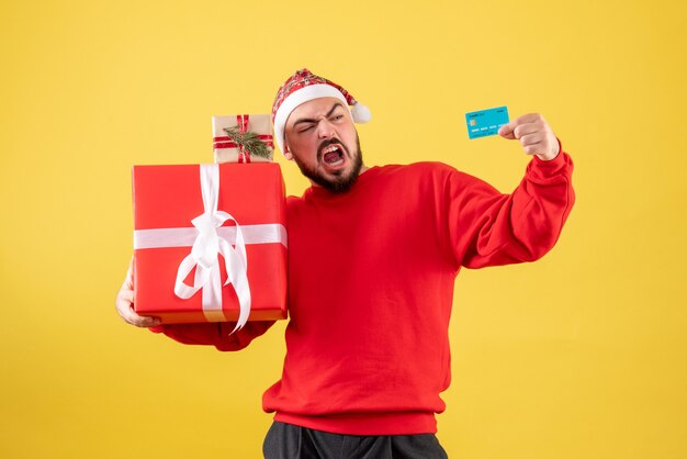 Vorderansicht junger Mann, der Weihnachtsgeschenk und Bankkarte auf gelbem Hintergrund hält