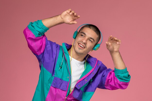 Vorderansicht junger Mann im bunten Mantel, der Musik mit Tanzbewegungen auf dem rosa Hintergrund hört