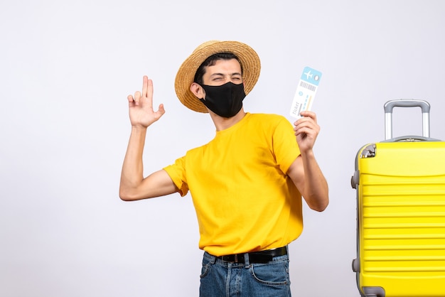 Vorderansicht junger Mann im gelben T-Shirt, das nahe gelbem Koffer steht, der Reiseticket hält, das Fingerpistole macht