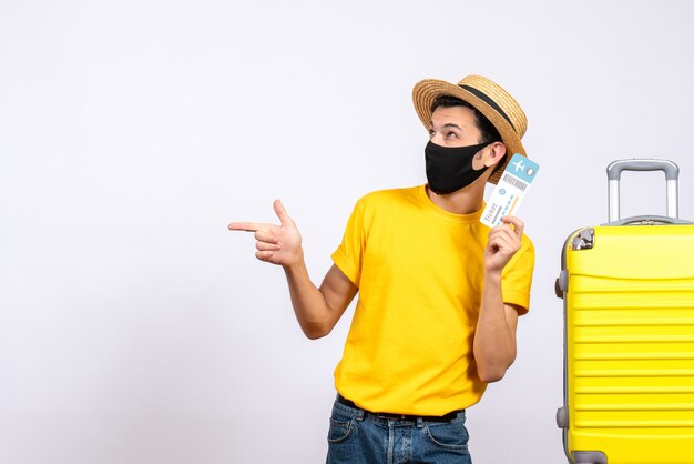 Vorderansicht junger Mann im gelben T-Shirt, der nahe gelbem Koffer steht und Reiseticket hält, das nach links zeigt