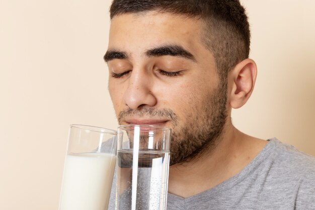 Vorderansicht junger Mann im grauen T-Shirt, das Gläser des Wassers und der Milch hält, die sie auf Beige riechen