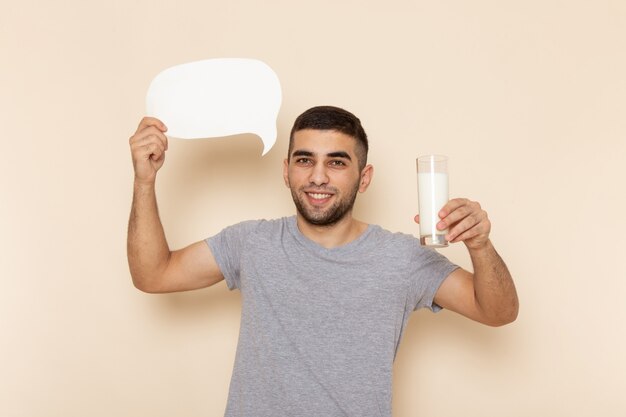 Vorderansicht junger Mann im grauen T-Shirt, das Glas Milch und weißes Zeichen auf Beige hält