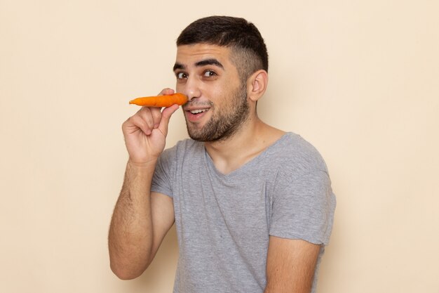 Vorderansicht junger Mann im grauen T-Shirt, das orange Karotte auf Beige hält