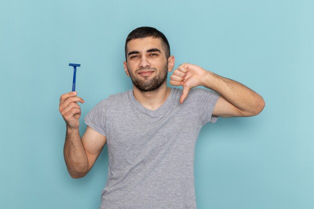 Vorderansicht junger Mann im grauen T-Shirt, das Rasiermesser hält und unähnliches Zeichen auf dem Blau zeigt