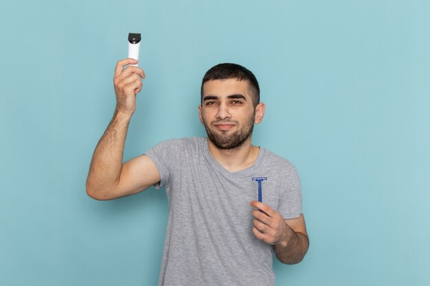 Vorderansicht junger Mann im grauen T-Shirt, das Rasiermesser und Elektrorasierer auf dem Blau hält