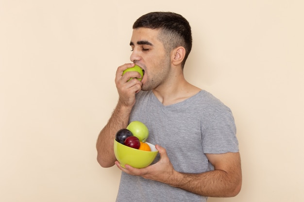 Vorderansicht junger Mann im grauen T-Shirt, der Platte mit Früchten beißt, die Apfel auf Beige beißen