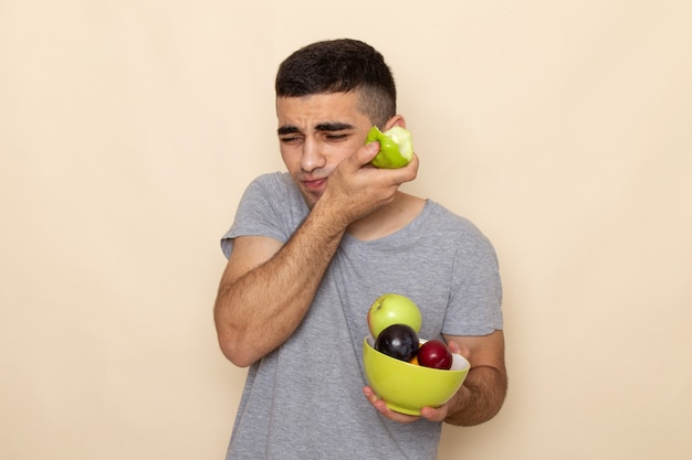 Vorderansicht junger Mann im grauen T-Shirt, der Platte mit Früchten beißt, die Apfel beißend Zahnschmerzen auf Beige haben