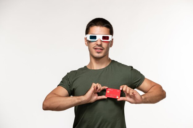 Vorderansicht junger Mann im grünen T-Shirt, das rote Bankkarte in -d Sonnenbrille auf hellweißem Wandfilm einsames Kino hält