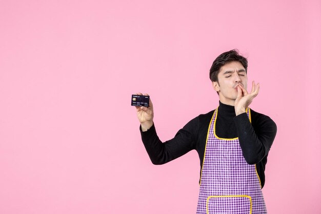 Vorderansicht junger Mann im Umhang mit Bankkarte auf rosafarbenem Hintergrund horizontale Farbe einheitlicher Job Kochen Arbeiter Küchenchef