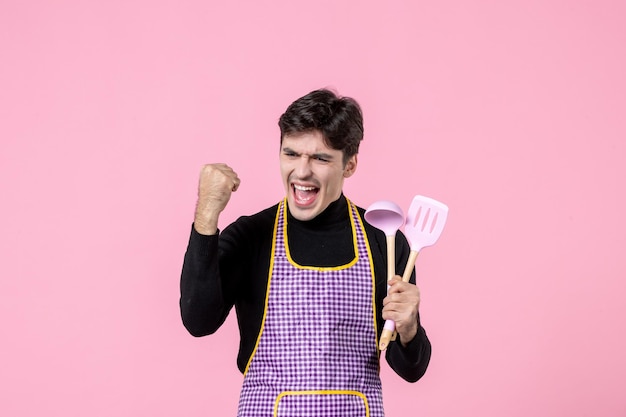 Vorderansicht junger Mann im Umhang mit Löffeln auf rosa Hintergrund Beruf Essen kochen Mahlzeit horizontale Farbe einheitliche Teigarbeit