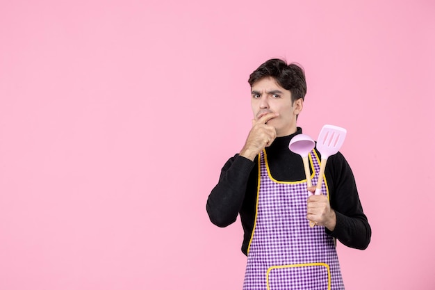 Vorderansicht junger Mann im Umhang mit Löffeln auf rosa Hintergrund Job Teig Beruf Küche einheitliche horizontale Kocharbeiter Cheffarbe