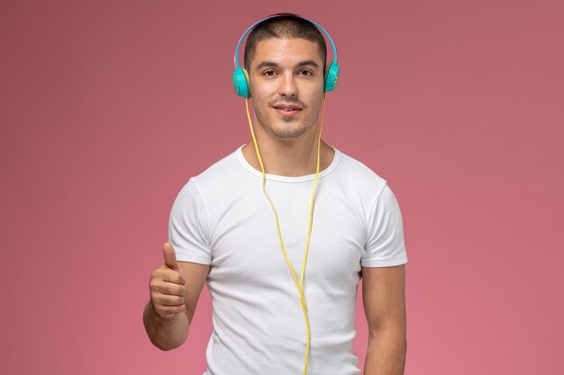 Vorderansicht junger Mann im weißen T-Shirt, das Musik über Kopfhörer auf dem hellrosa Schreibtisch hört