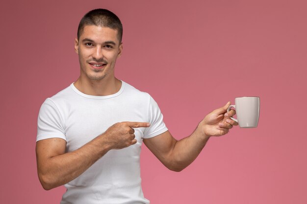 Vorderansicht junger Mann im weißen T-Shirt, das Tasse mit Kaffee auf dem rosa Hintergrund hält