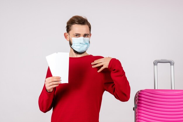 Vorderansicht junger Mann in der sterilen Maske, die Tickets auf Flug der weißen Wandreise Covid-Trip-Urlaubs-Emotionsvirus hält
