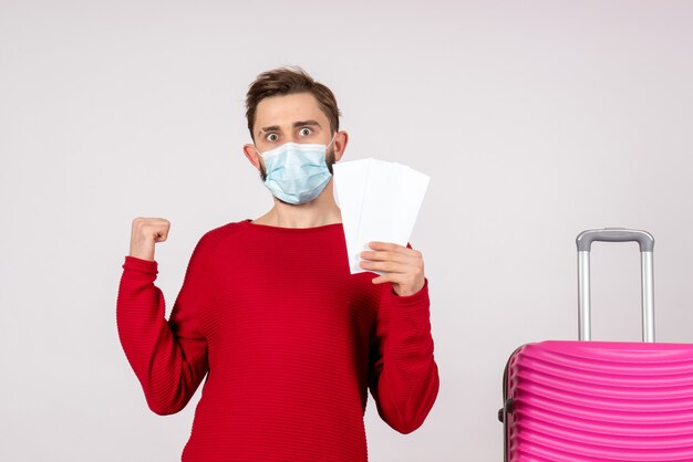 Kostenloses Foto vorderansicht junger mann in der sterilen maske, die tickets auf weißer wand covid-plane holiday emotion virus flugfarbe hält