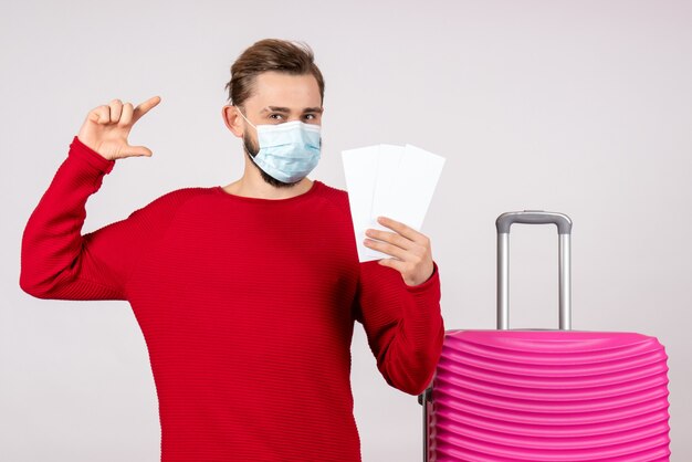 Vorderansicht junger Mann in der sterilen Maske, die Tickets auf Weißwand-Reise-Covid-Flugreise-Urlaubsfarbemotionsvirus hält