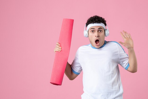 Vorderansicht junger Mann in Sportkleidung mit Yogamatte und Kopfhörern auf rosa Wand