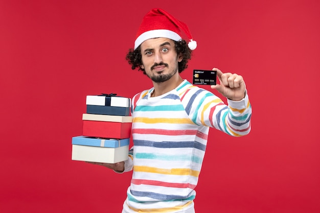 Vorderansicht junger Mann mit Feiertagsgeschenken und Bankkarte auf rotem Schreibtisch Neujahrsgeldrot