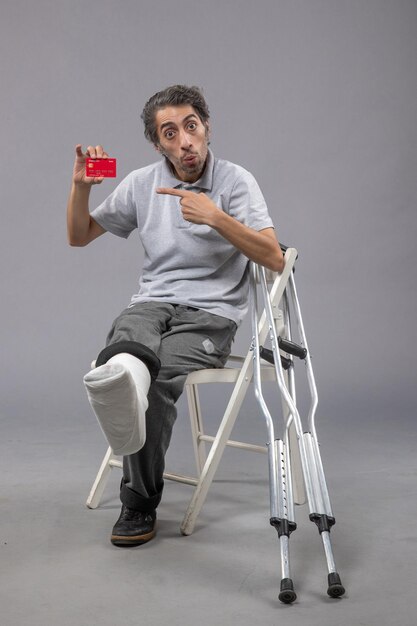 Vorderansicht junger Mann mit gebrochenem Fuß und Verband, der Bankkarte auf grauem Bodenunfall hält, verdreht Beinfuß menschlicher Schmerz