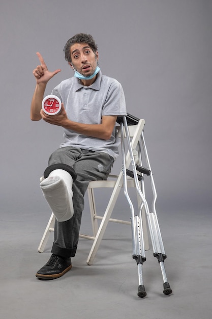 Vorderansicht junger Mann mit gebrochenem Fuß und Verband, der eine rote Uhr an der grauen Wand hält
