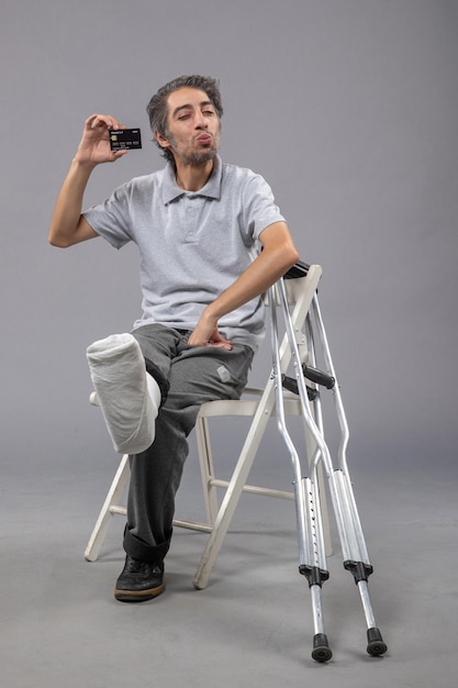 Vorderansicht junger Mann mit gebrochenem Fuß und Verband, der schwarze Bankkarte auf der grauen Wand hält menschlicher Twist männlicher Unfallfußschmerz